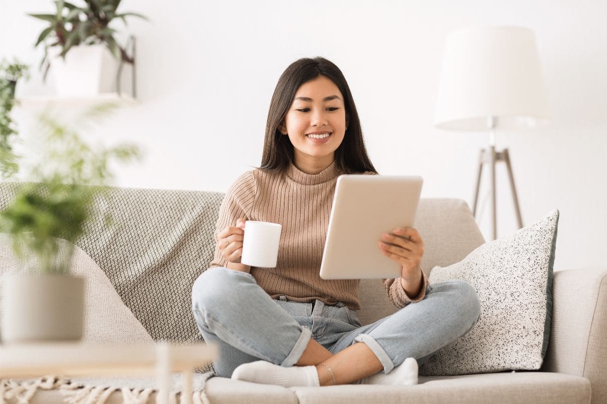 A woman sitting on a couch with a tablet and coffee cup, engrossed in her digital device and enjoying a warm beverage.